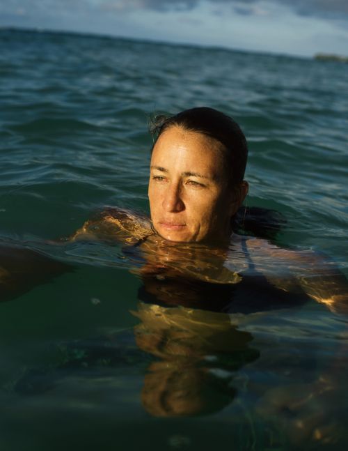 Close-up of a woman swimming in the ocean. She is partially submerged in the water, gazing off to the side. Waves and sky are visible.