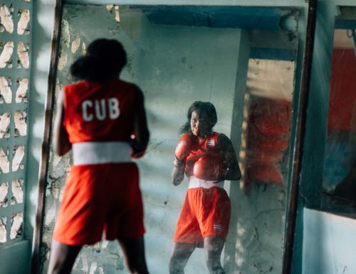 Woman boxer in red uniform with 'CUB' on back, reflected in a mirror, prepares in a boxing ring, showcasing focus and determination.