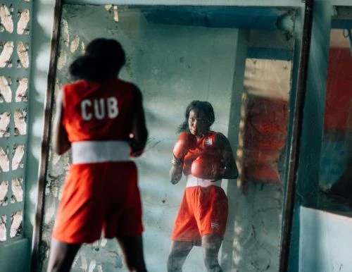 Woman boxer in red uniform with 'CUB' on back, reflected in a mirror, prepares in a boxing ring, showcasing focus and determination.