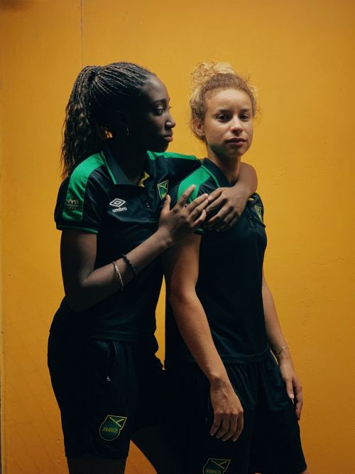 Two women soccer players in black and green Jamaica uniforms pose against a yellow wall, one with an arm around the other.