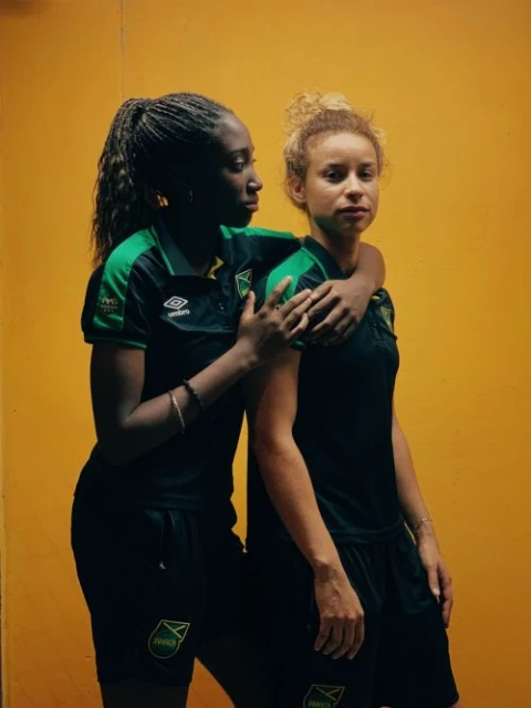Two women soccer players in black and green Jamaica uniforms pose against a yellow wall, one with an arm around the other.