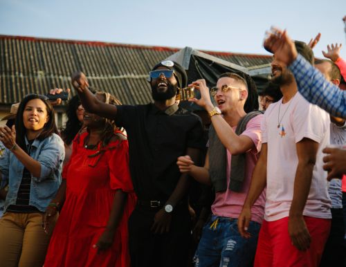 Group of people gathered outdoors, some holding up phones, and cheering, in casual clothing, against a background with a building structure.