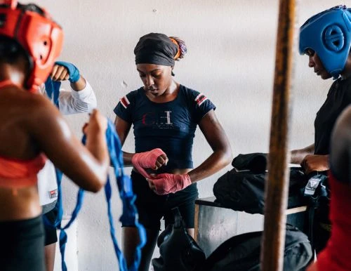 Three female boxers prepare in gym: one wraps pink hand wraps, another holds blue tape, and a third wears a blue head guard.