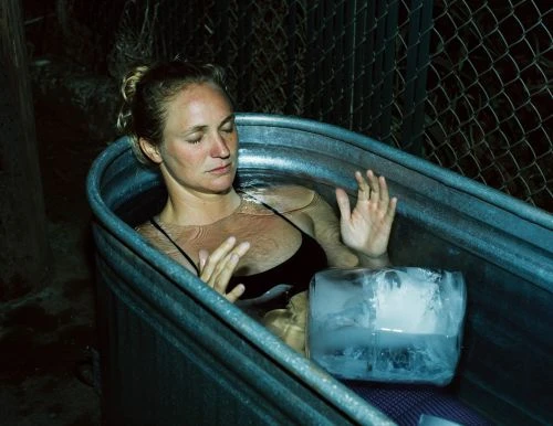 Adult female immerses in an ice bath outdoors with a large ice block, eyes closed, wearing a black top, inside metal container.