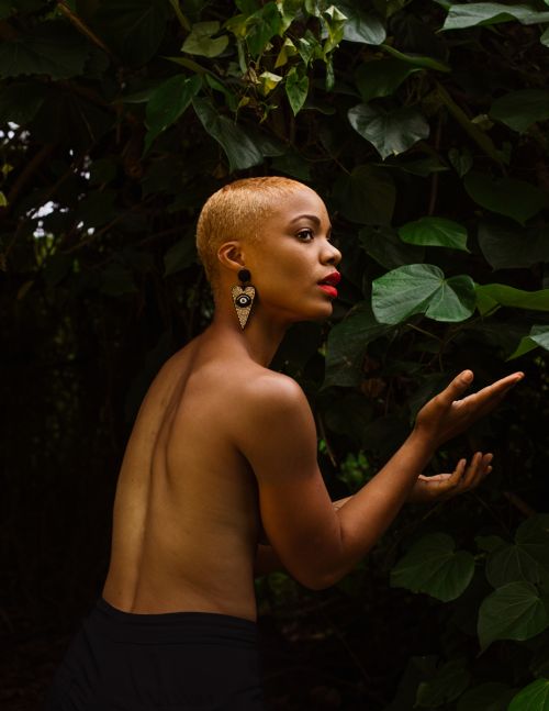 Portrait of a short-haired woman in an outdoor setting, adorned with statement earrings and bold red lipstick, set against green leaves.