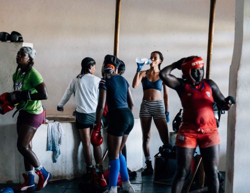 Group of female boxers in workout gear at boxing gym. One woman drinks from a water bottle in the gym; others wear headgear and gloves.