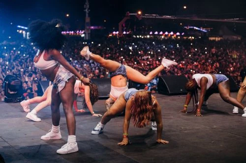 Energetic image of female dancers performing on stage at an outdoor music festival with a large crowd, bathed in vibrant stage lighting.
