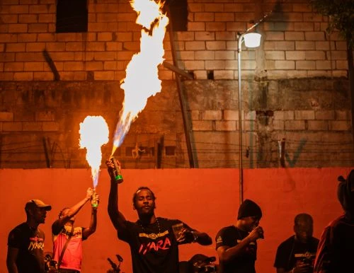 A group of men are performing with flames in an urban setting at night, creating a dramatic visual display with fire and light.