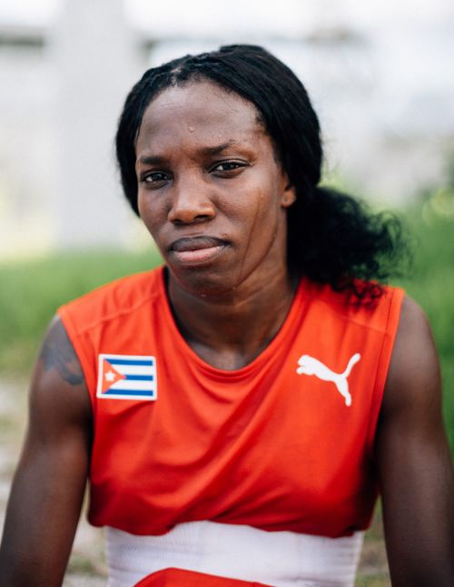 Adult female athlete in red tank top with Cuban flag and Puma logo, outdoor medium shot, with blurred green foliage background.