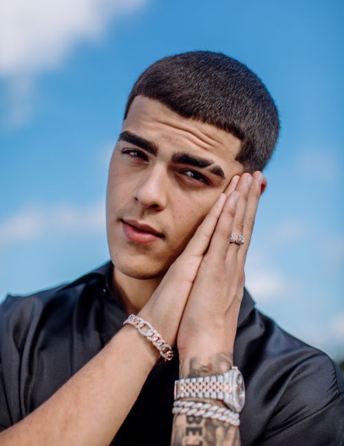 Close-up portrait of a young adult male outdoors against a bright blue sky, wearing jewelry, with his hands clasped. 