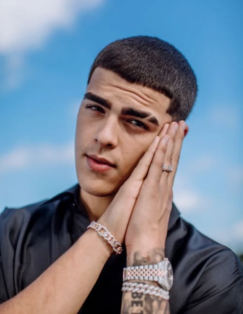 Close-up portrait of a young adult male outdoors against a bright blue sky, wearing jewelry, with his hands clasped. 
