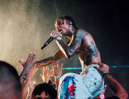 Shirtless male rapper on shoulders performing on stage, holding microphone with raised hands in crowd, concert with dark background.