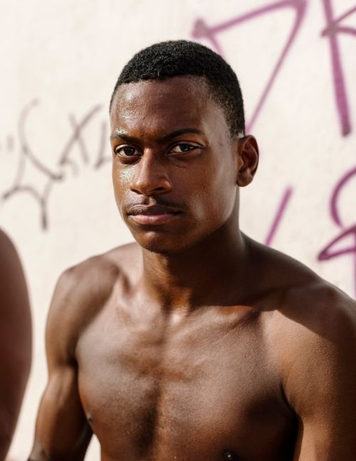 Portrait of a shirtless young Black man with short dark hair standing against a weathered wall with graffiti