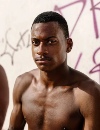 Portrait of a shirtless young Black man with short dark hair standing against a weathered wall with graffiti