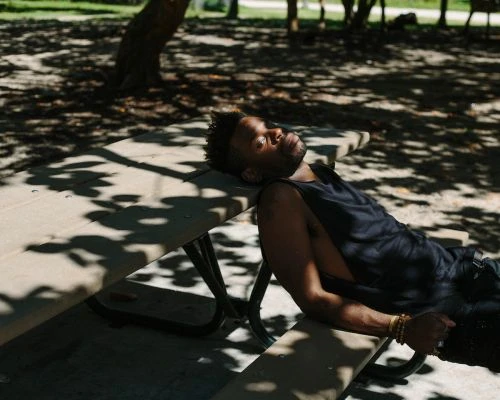 A black man relaxes on a picnic table in a sun-dappled park, eyes closed, wearing a black tank top, bracelets, and dark pants, enjoying the peaceful outdoor setting.