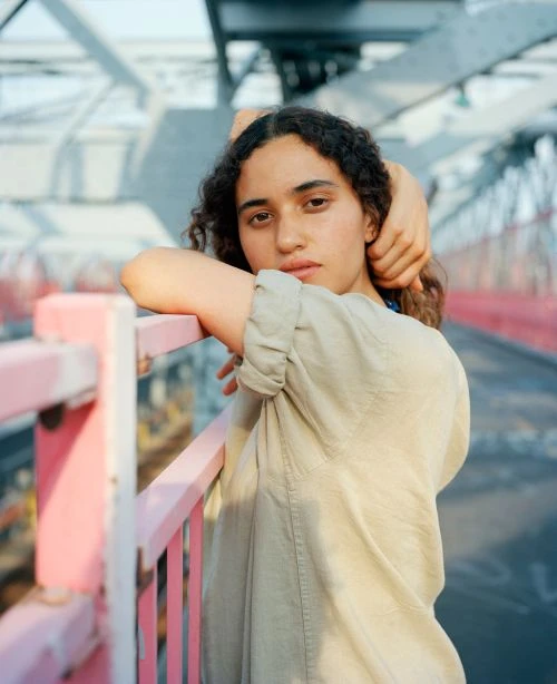 Young woman with curly hair leans against pink bridge railing in a medium shot on a sunny day, wearing a light-green shirt.