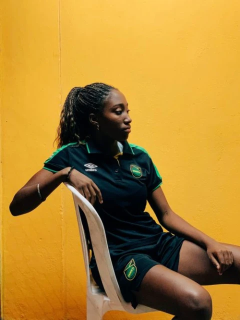 Young woman in a Jamaica soccer jersey poses against a yellow wall while seated on a white chair, looking away from the camera.