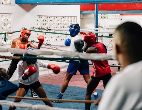 Female boxers sparring in a boxing ring wearing protective gear with an adult male observer in a boxing gym with red and blue trim.