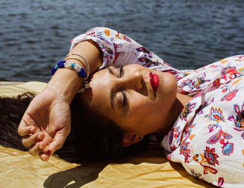 Woman with closed eyes in floral top relaxes on a gold fabric by the water in an eye-level, outdoor shot with natural lighting.