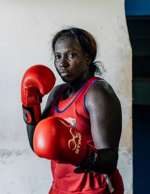 Portrait of a female boxer in red boxing gloves and jersey, standing in front of a textured white wall with a concentrated expression.