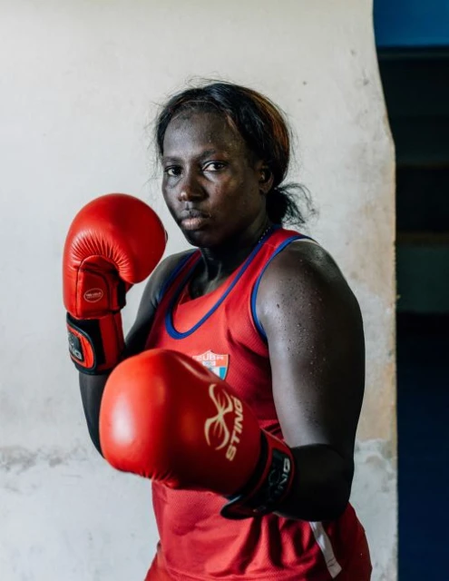 Portrait of a female boxer in red boxing gloves and jersey, standing in front of a textured white wall with a concentrated expression.