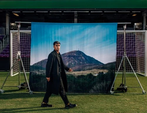 Man in a black coat walking on a soccer field with a mountain backdrop, showcasing a fashion editorial on an outdoor sports venue.