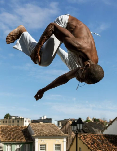 An athletic African-American man performs an acrobatic backflip with a city building backdrop and a clear blue sky in an outdoor setting.