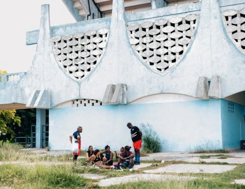 Group of athletes in sportswear sit on the grass near a stadium; some stand and chat outdoors during daylight on a sunny day.