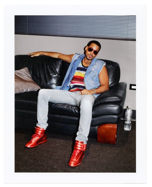 Man wearing sunglasses, denim vest, striped shirt, light jeans, and red high-top sneakers seated on a black leather sofa.