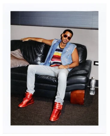 Man wearing sunglasses, denim vest, striped shirt, light jeans, and red high-top sneakers seated on a black leather sofa.
