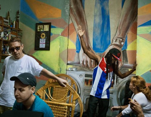 An indoor candid shot shows a graffiti-covered wall backdrop, featuring three adults, one wearing a Cuban flag tank top, and one with sunglasses.