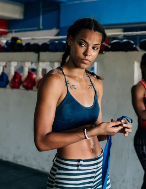 Athletic woman in workout clothes wraps blue boxing hand wraps in a gymnasium with equipment visible. Fitness and healthy lifestyle.