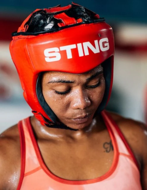 Close-up of a female boxer wearing a red headgear with the word 'STING' in white letters, looking down with a serious expression.