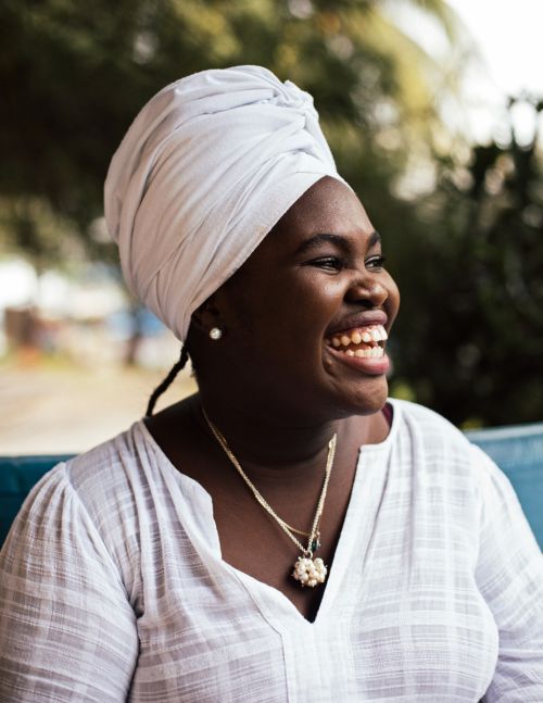Close-up of a smiling adult woman with dark skin wearing a white headscarf and a white shirt, captured outdoors with soft, natural lighting.