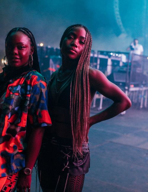 Two women pose in an outdoor concert venue with blue lighting and stage equipment visible in the background, creating a vibrant atmosphere.