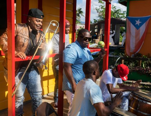 Five musicians are playing instruments outside a building with a Puerto Rican flag. One man plays the trombone and others drums.