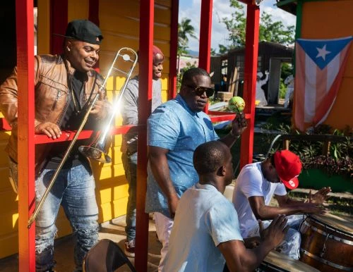 Five musicians are playing instruments outside a building with a Puerto Rican flag. One man plays the trombone and others drums.