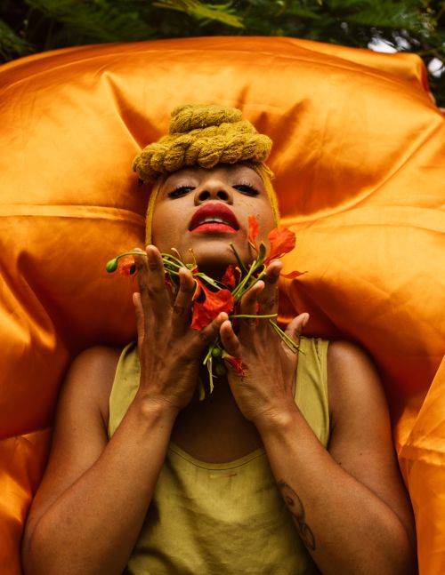 Eye-level portrait of an adult female lying on gold satin fabric, surrounded by green foliage, while holding red flowers to her face.