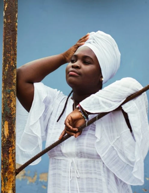 Portrait of a Black woman with a white headscarf, resting her arm on a rusty metal bar against a textured blue wall background.