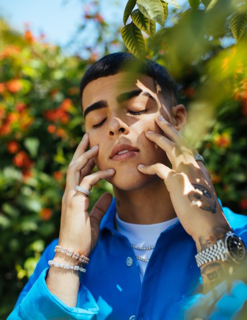 Close-up portrait of a young man wearing a vibrant blue shirt and jewelry with eyes closed outdoors among foliage