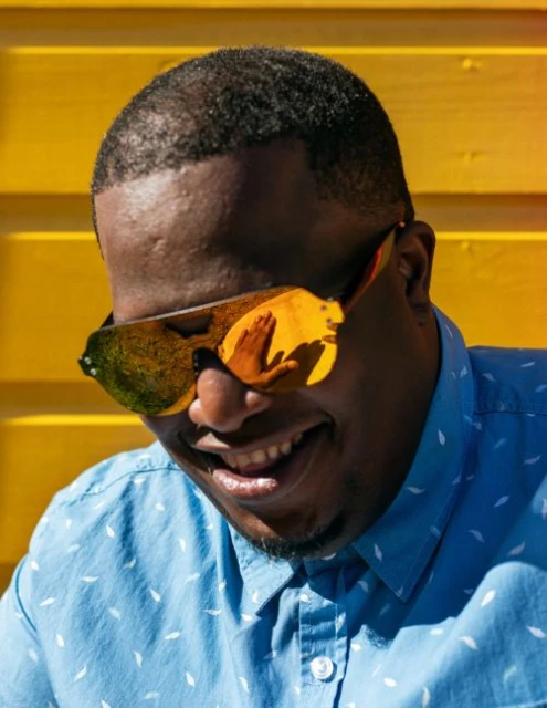 Person smiles in a headshot, wearing reflective sunglasses, a patterned shirt, against a bright yellow background on a sunny day.