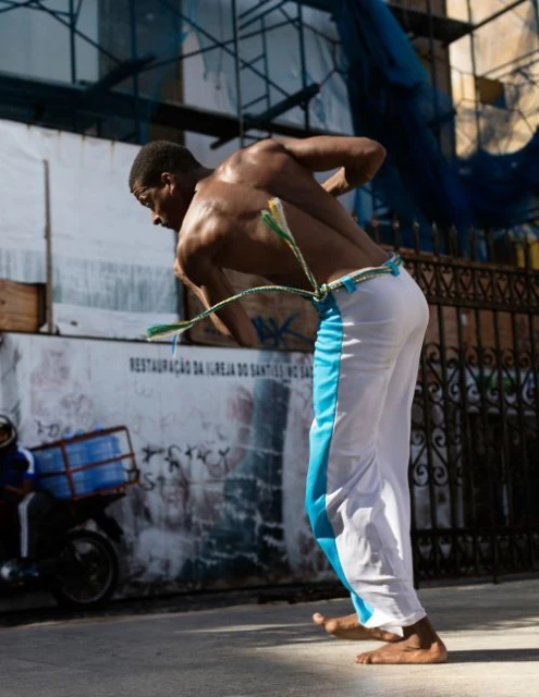 An African American man wearing white pants is practicing Capoeira on a city street in front of a building with a scooter nearby.