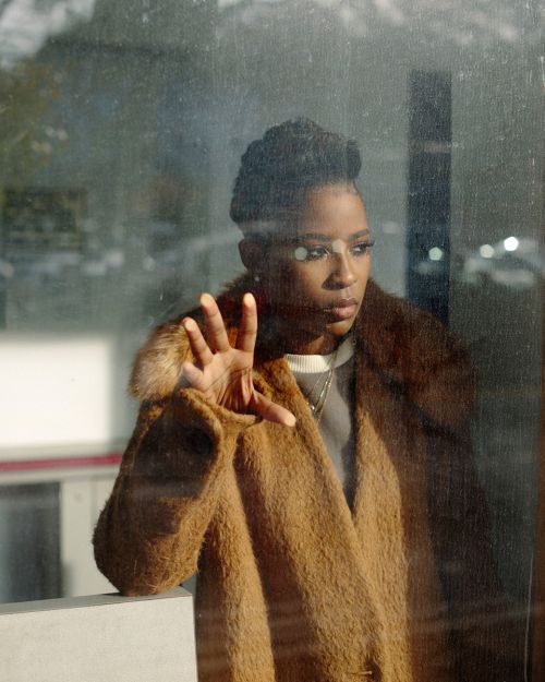 Close-up portrait of a young woman with dark hair wearing a black turtleneck, against a blurred, warm-toned background in a studio setting.