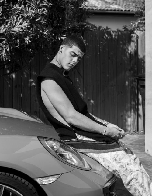 A black and white portrait of a young adult man posing next to a luxury sportscar outdoors with a fence and foliage background.
