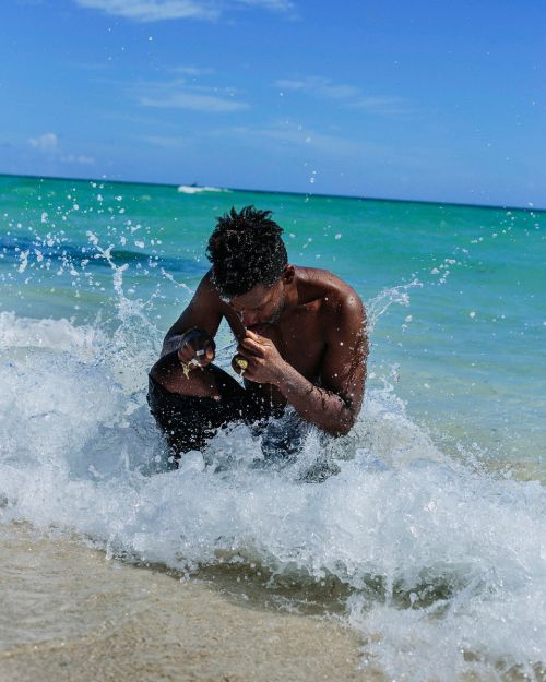 Person sits in ocean surf at beach, close-up medium shot. Water splashes around the individual against blue sky and turquoise water backdrop.