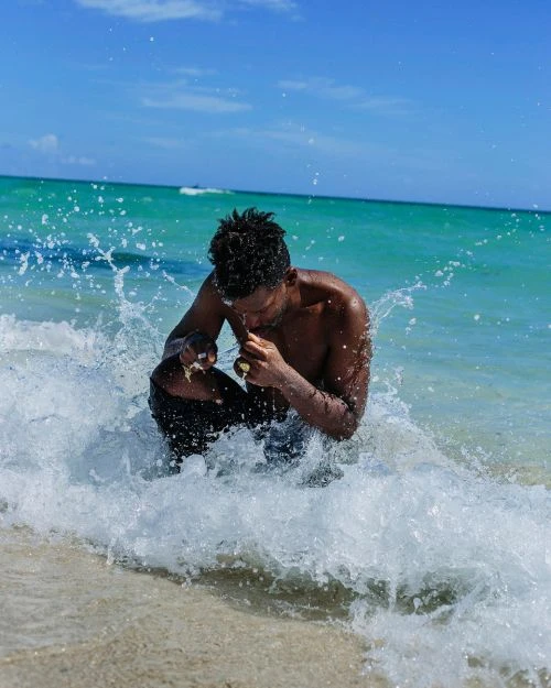Person sits in ocean surf at beach, close-up medium shot. Water splashes around the individual against blue sky and turquoise water backdrop.