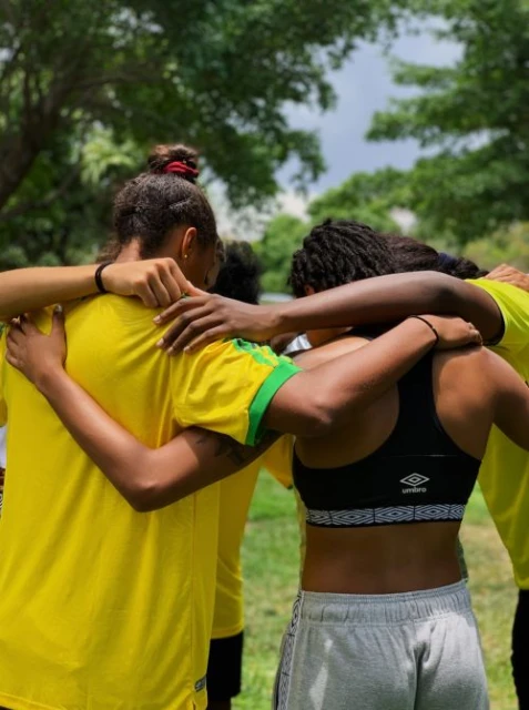 Outdoor photo of a group of young adult women in sports attire huddling together with their arms around each other against a blurred backdrop.