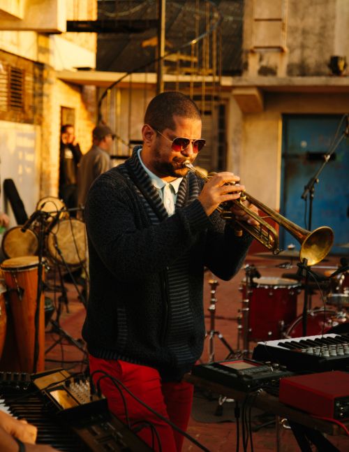 Musician in a knit cardigan and sunglasses plays a trumpet at an outdoor music venue, with other musicians and instruments visible.