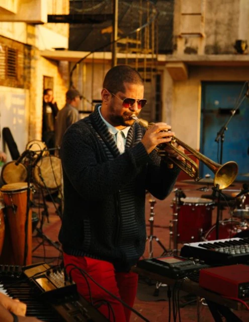 Musician in a knit cardigan and sunglasses plays a trumpet at an outdoor music venue, with other musicians and instruments visible.