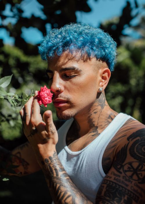Close-up portrait of a tattooed man with bright blue curly hair holding a small red rose to his nose outdoors.